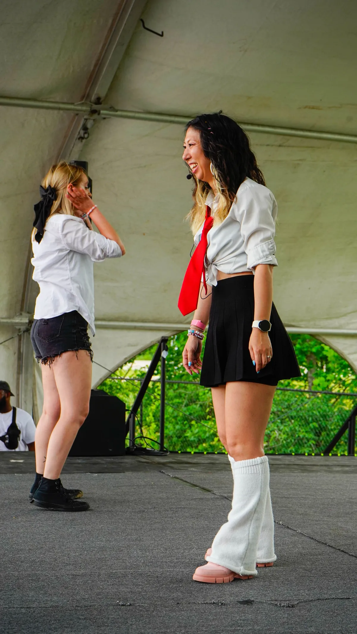 A K-Pop dance instructor wearing a headset microphone, a tied white shirt with a red tie, black skirt and white leg warmers laughs mid-lesson on a tented outdoor stage at Knox Pop Con.